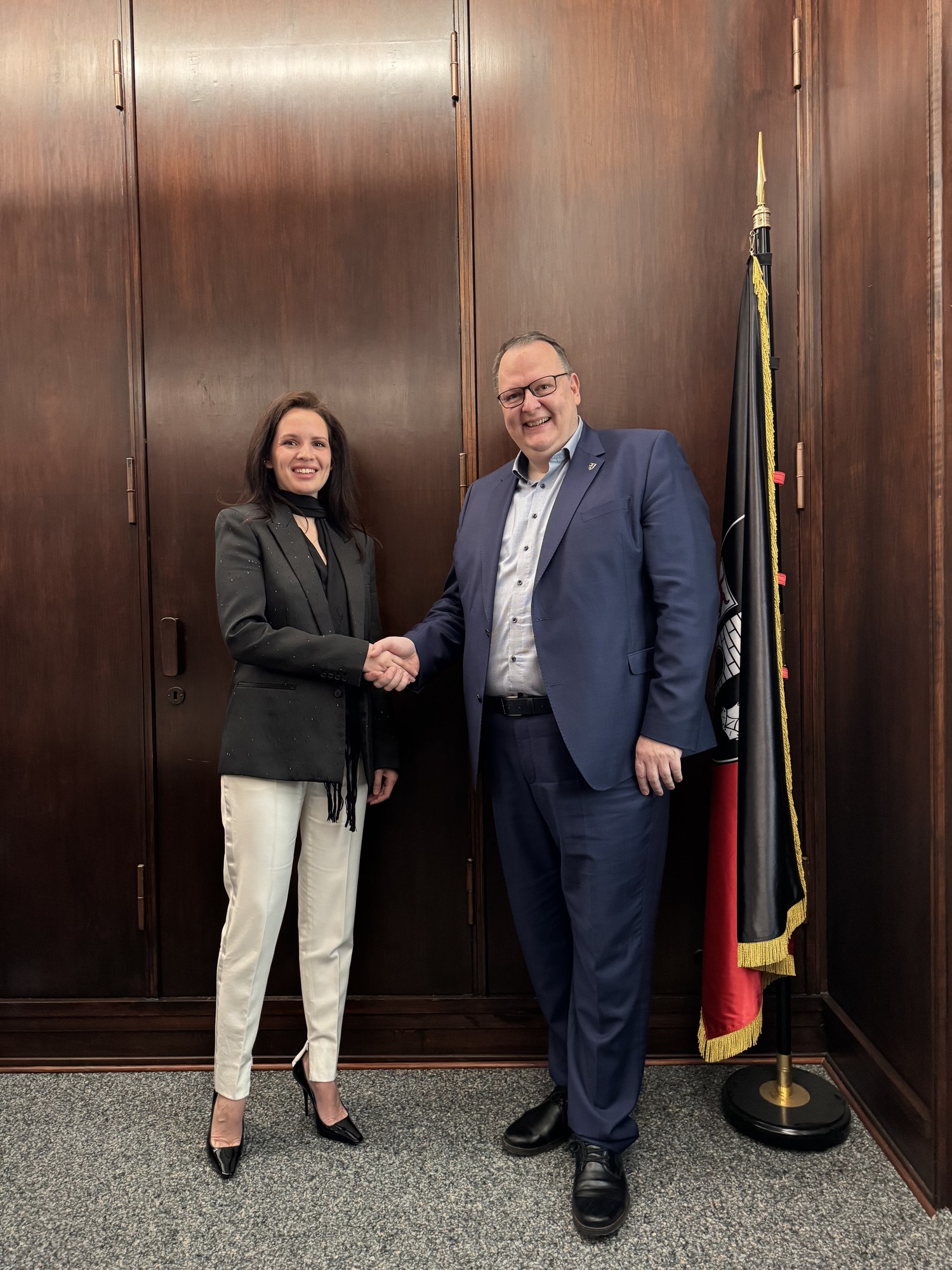 Anna Bon shaking hands with Bürgermeister Heiko Breithaupt in Blankenburg town hall, the city flag beside them