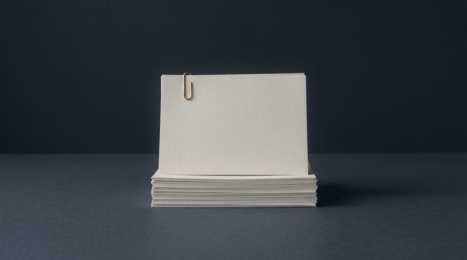 A squared stack of pale paper on a dark ground, one brass clip on the top sheet